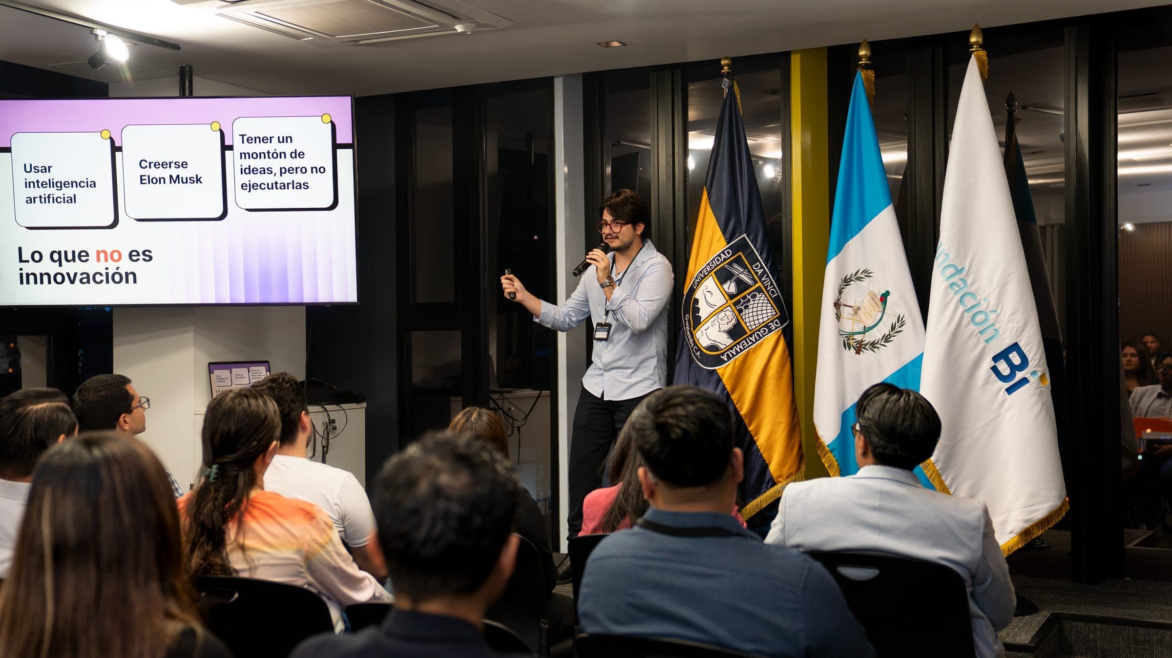 Marcello Sisniega presentando ante una audiencia junto a banderas institucionales en un evento con Fundación BI